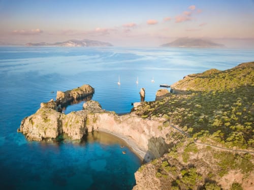 Scogliere costiere e acque turchesi dell'Hotel Principe di Rinella a Rinella, Italia con barche a vela e isole in lontananza