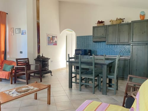 Open-plan kitchen and dining area in Orosei villa with green cabinetry, blue tile backsplash, dining table, and wood stove