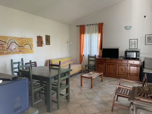 Bright living room in Orosei villa with dining table, sofa, TV, and orange curtains overlooking sea view