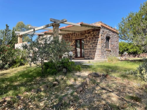 Villa in pietra a Orosei con pergolato in legno, zona giardino con sedute e ulivi sotto cielo azzurro