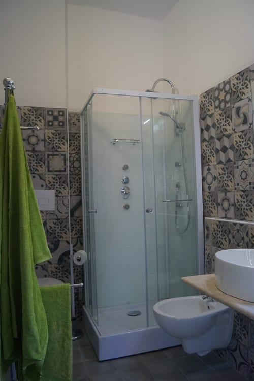 Bagno moderno della villa di Orosei con cabina doccia in vetro, sanitari bianchi e pareti rivestite di piastrelle decorative con asciugamano verde
