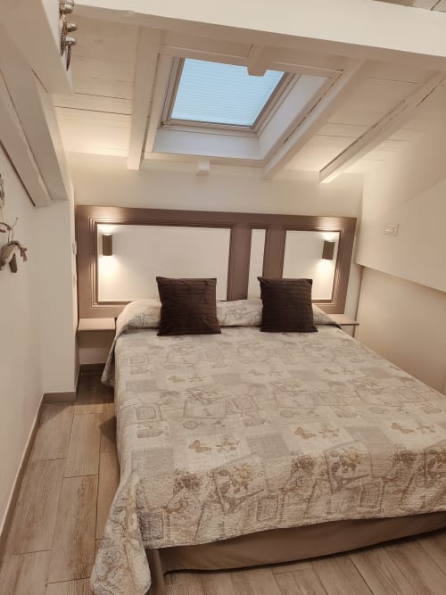 Moderno loft camera a Ventimiglia con lucernario, letto matrimoniale, cuscini scuri, pavimento in legno e testata specchiata.