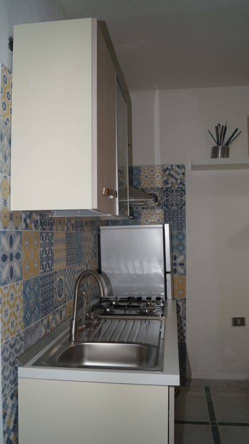 Compact kitchen in Orosei apartment with patterned blue and yellow tile backsplash, gas cooktop, sink, and white cabinetry