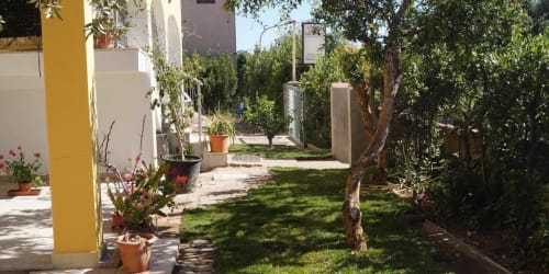 Sunny garden courtyard of Sa Domo De Mannoi apartment in Orosei with lush greenery, yellow column, potted flowers, and paved
