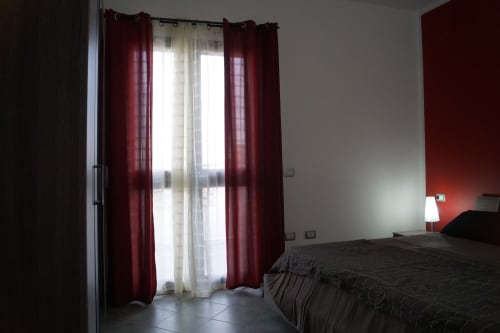 Modernes Schlafzimmer in der Wohnung von Orosei mit bordeauxfarbenen Vorhängen, Glasschiebetüren zur Veranda und dunkler Bettwäsche
