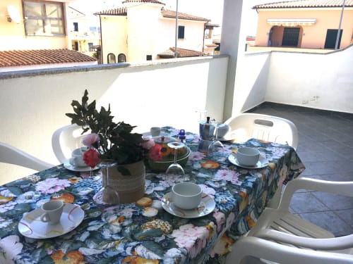 Außenveranda mit Blumentischtuch, Gedeck, Blumentöpfen und weißen Stühlen mit Blick auf das Dorf Orosei