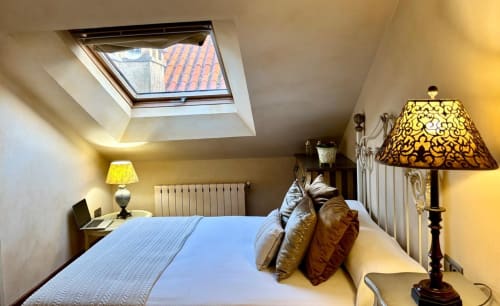 Luminoso dormitorio abuhardillado en Baiona con claraboya, ropa de cama azul, almohadas decorativas, lámparas mesita y vista al mar desde ventana de techo