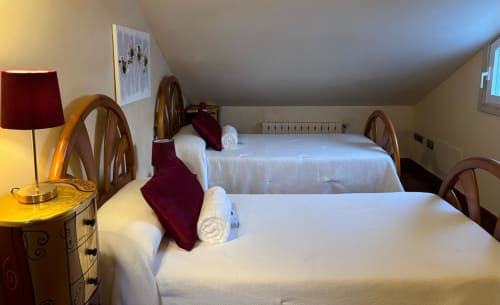 Luminoso dormitorio abuhardillado en Holibai, Baiona con dos camas, cabeceros de madera, detalles burdeos, lámpara y vistas al mar