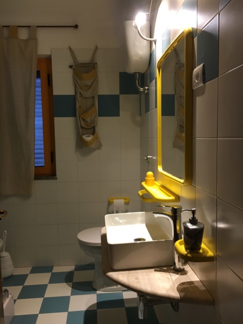 Modern bathroom in Orosei apartment with white sink, yellow-framed mirror, teal and white checkered floor, and shower area.