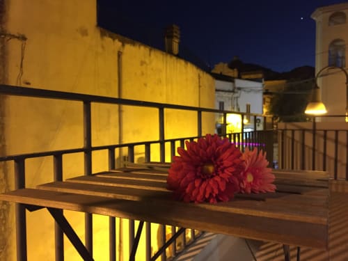 Red flowers on wooden terrace at night, illuminated historic buildings in Orosei, Italy