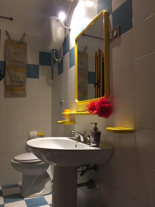 Modern bathroom with pedestal sink, yellow-framed mirror, red flowers, and blue-white tiled walls in Orosei apartment