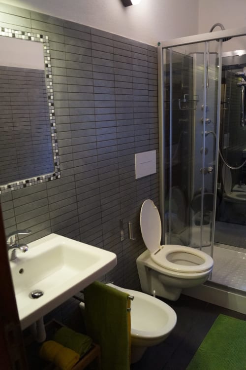 Modern bathroom with dark tile walls, white fixtures, and glass shower enclosure at Loddo Paola apartment in Orosei