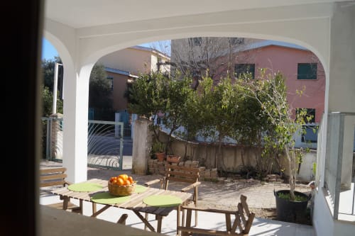 Sunny veranda with wooden dining table at Orosei apartment, featuring potted plants and red-brick buildings