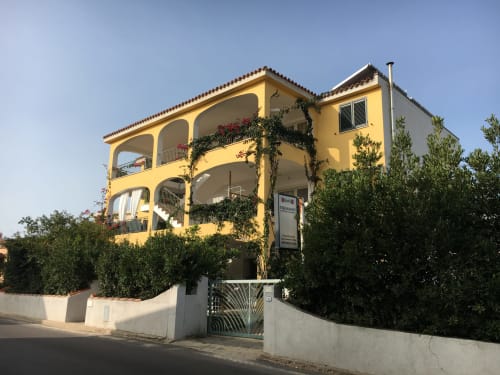 Bright yellow Mediterranean villa in Orosei with curved balconies, ivy-draped walls, and white gate surrounded by green