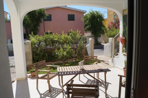 Sunny courtyard with wooden seating, lush plants, and pink building in Orosei apartment rental