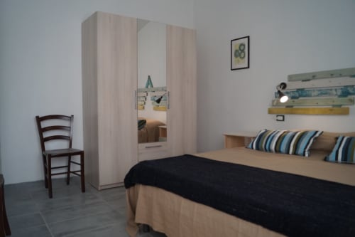 Bright bedroom in Orosei apartment with wooden wardrobe, striped pillows, dark bedcover, and chair