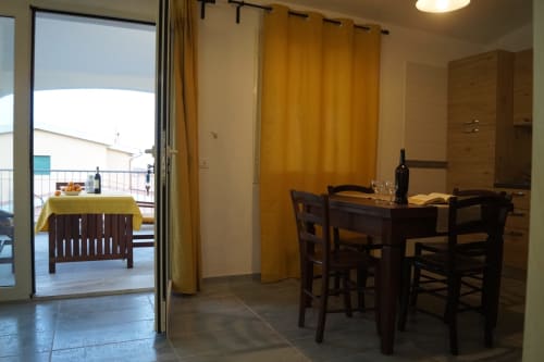 Dining area with dark wood table and chairs, yellow curtains, and open door to veranda in Orosei apartment