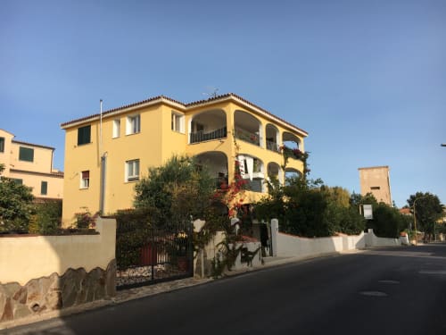 Bright yellow apartment building in Orosei, Italy with arched balconies, surrounded by gardens and white walls