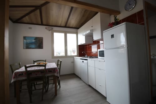Kitchen-dining area with white appliances, red tile backsplash, wooden table with chairs, and city view windows in Orosei
