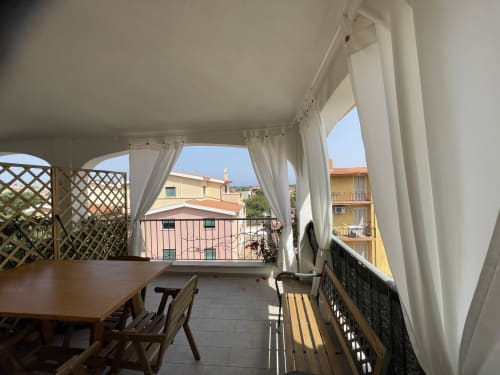 Covered veranda with white curtains overlooking Orosei, featuring wooden dining table and armchairs