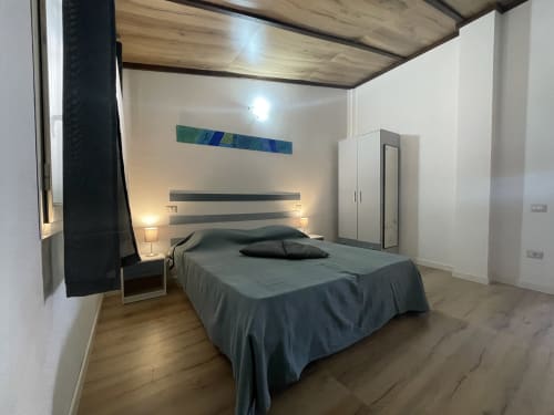 Modern bedroom with gray bed, wooden ceiling, striped headboard with wall art, wardrobe, and side table in Orosei apartment