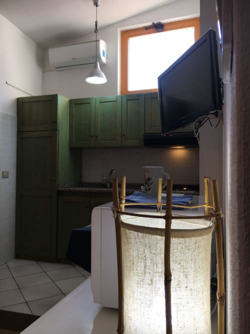 Cucina verde con piano di lavoro scuro, TV a parete, lampada a sospensione e cesto per il bucato nella stanza Orosei