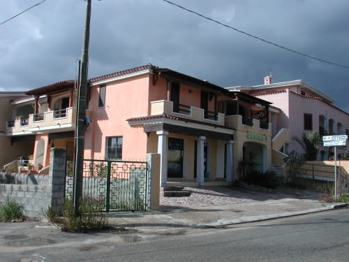 Domo de Mannoi apartment in Orosei with terracotta-tiled roof, pink and beige facade, and outdoor veranda under cloudy sky