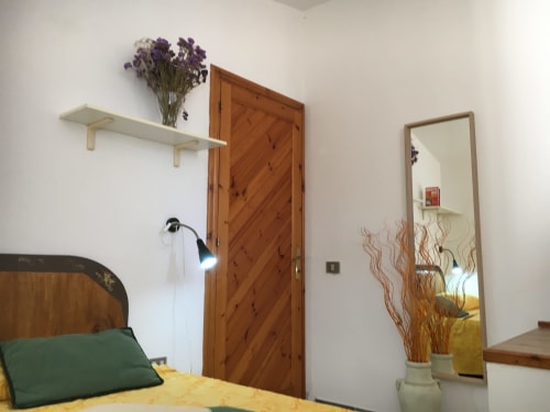 Camera da letto in Casa Arborea A, Orosei. Porta in legno, scaffali bianchi con fiori secchi, specchio, cuscino verde sul letto.