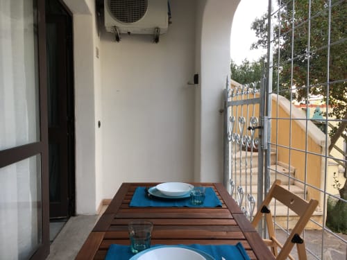 Outdoor veranda dining area in Orosei apartment with wooden table, place settings, and metal gate overlooking yellow building