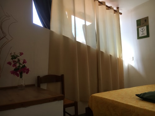 Stanza luminosa a Casa Arborea A a Orosei con tende color crema, arredi in legno, fiori rosa e luce naturale abbondante