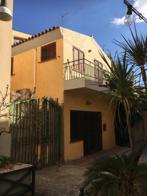 Bright yellow Mediterranean apartment building in Orosei with balcony, palm trees, and metal gate entrance