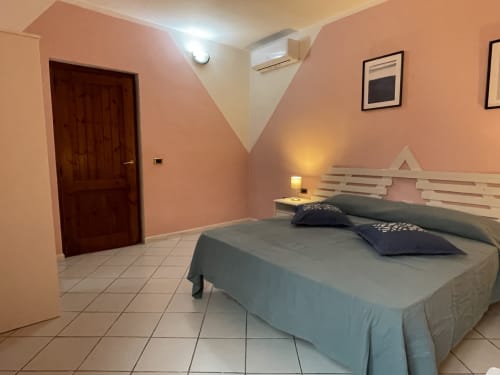Modern bedroom in Orosei apartment with pink walls, queen bed, dark wood door, and air conditioning unit