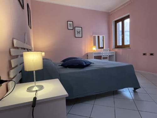 Modern bedroom at Loddo Paola in Orosei with pink walls, dark blue bed, desk lamp, and city views through windows