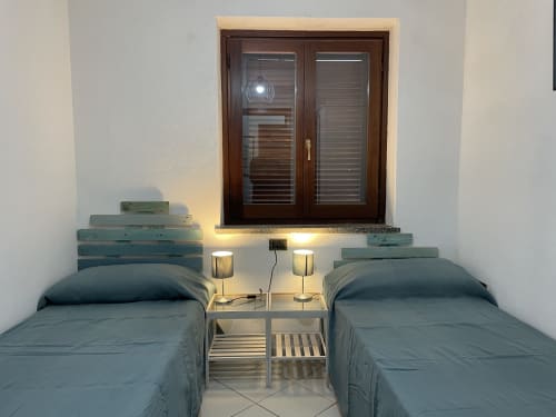 Twin bedroom with bedside table and lamps, wooden doors with shutters opening to veranda in Orosei apartment