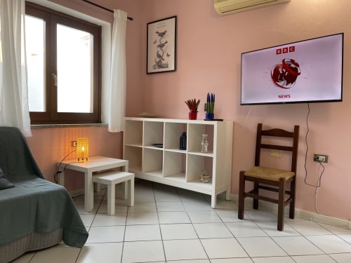 Modern living room in Orosei apartment with pink walls, TV, shelving unit, seating, and potted plants