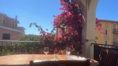 Sunny veranda at Orosei apartment with vibrant magenta bougainvillea flowers, wooden table, and wine glasses overlooking city