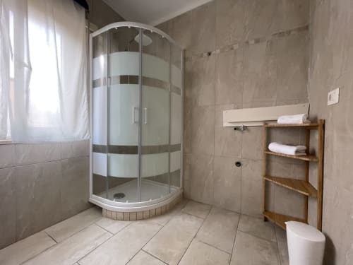 Modern bathroom with curved glass shower enclosure, wooden shelf, and beige tile flooring in Orosei apartment