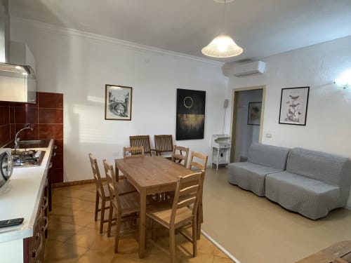 Open-plan apartment in Orosei with wooden dining table, kitchen, grey sofa, and framed artwork on white walls