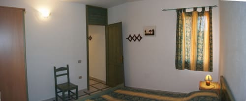 Camera Casa Arborea G di Orosei con porte verdi, letti, poltroncine, lampada, finestra con tende e arredamento rustico