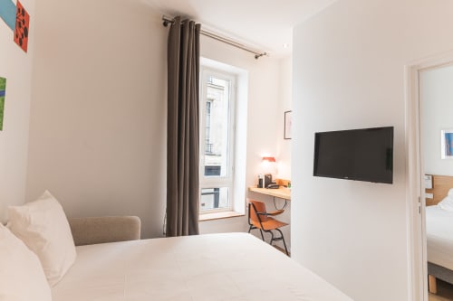 Suite moderne à l'Hôtel Louvre Richelieu Paris avec lit blanc, TV murale, bureau de travail et fenêtre aux rideaux gris