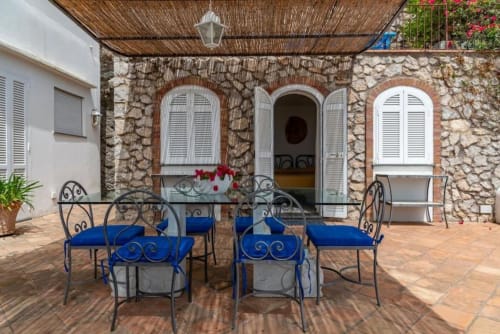 Terrazza all'aperto di La Conciergerie Capri con sedie in ferro battuto blu, edificio in pietra e finestre ad arco