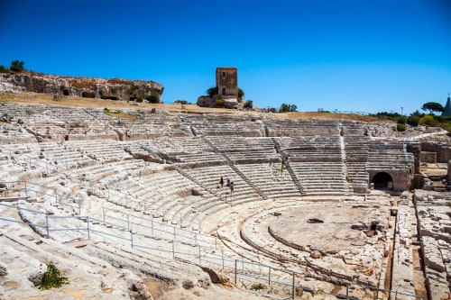 Rovine dell'antico anfiteatro greco a Siracusa con gradinate in pietra e torre, cielo azzurro limpido