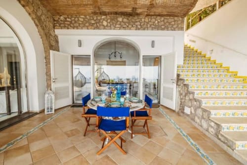 Elegante sala da pranzo di villa a Capri con vista sul mare, sedie blu al tavolo rotondo, arcata in pietra e scala decorativa