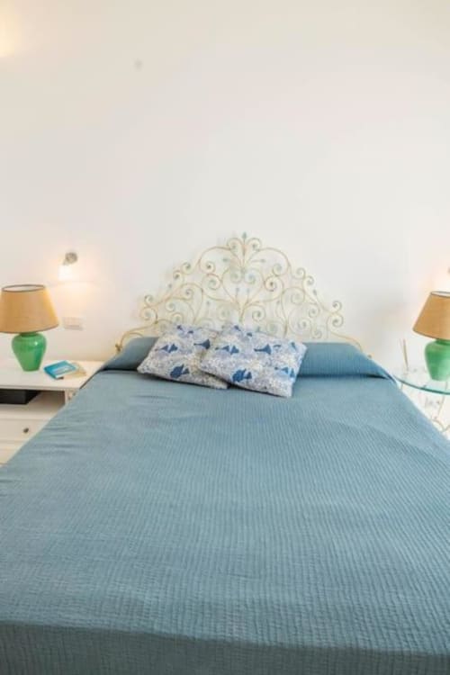 Camera luminosa di Villa La Conciergerie Capri con testata ornamentale, biancheria azzurra, lampade verdi e arredamento costiero