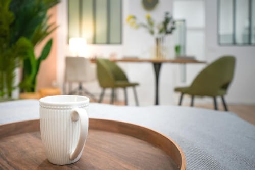 White ribbed mug on wooden tray in Wellkhome apartment lounge, Bordeaux, with dining area and green chairs blurred behind