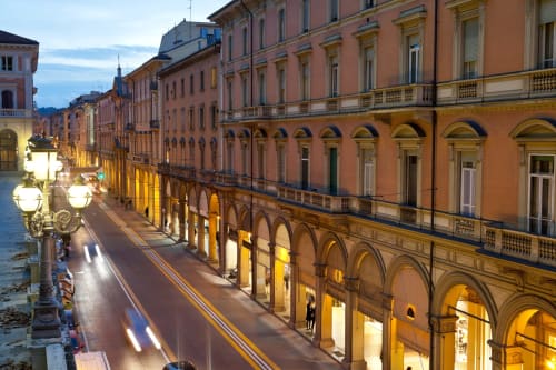 Via porticata storica a Bologna al tramonto con negozi illuminati e edifici in terracotta
