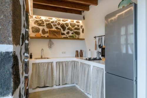 Rustic kitchen in El Granero cottage at Finca de La Geria with stone wall, wooden beams, and cream curtains.