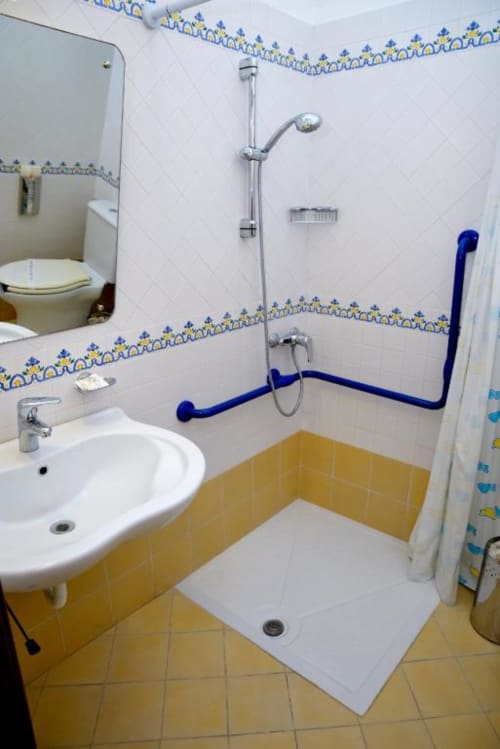 Accessible bathroom at Hotel Principe di Rinella with white and yellow tiles, blue grab bars, walk-in shower, and sink with