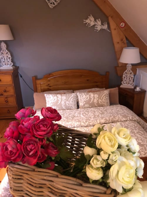 Chambre au Brise de Lin au Clos Mahiel avec lit en bois, coussins décoratifs lin, roses fraîches dans un panier, tables de nuit avec lampes raffinées.