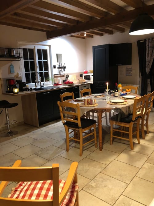 Spacieuse salle à manger du gite avec table ronde, chaises en bois, cuisine équipée, poutres apparentes et vue sur le jardin au Clos Mahiel